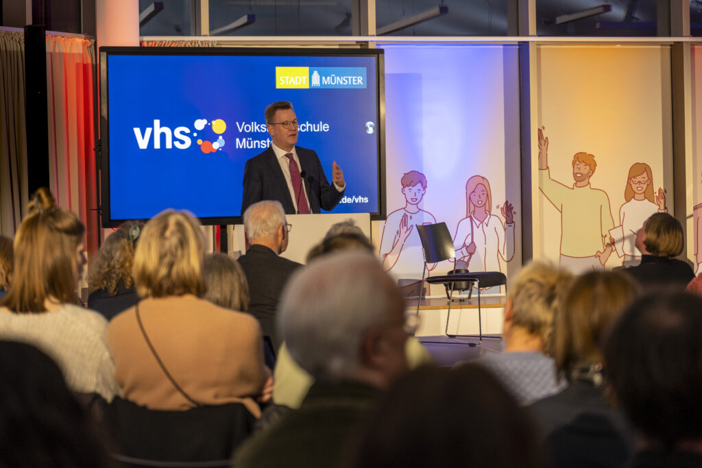 Prof. Dr. Johannes Wessels, Rektor der Uni Münster, am Rednerpult vor elektronischem Board mit vhs-Logo.